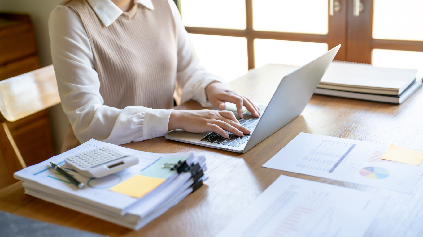 Mulher trabalhando em um laptop com documentos contábeis, planilhas e calculadora sobre a mesa, representando gestão financeira e contabilidade para pequenas empresas.